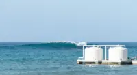 A large turquoise ocean wave rising far out at sea behind a small white coastal plant with two round tanks, under a clear blue sky