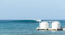 A large turquoise ocean wave rising far out at sea behind a small white coastal plant with two round tanks, under a clear blue sky