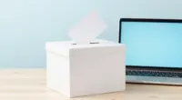 A plain white ballot box with a blank card in its slot on a light wooden desk, an open laptop with a blank pale blue screen beside it