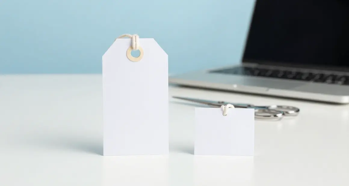 Two blank white paper price tags on a bright white desk, one large and one much smaller, with silver scissors and a laptop out of focus behind them