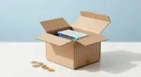An open cardboard shipping box on a bright white desk with a small glowing server inside and a few gold coins next to it