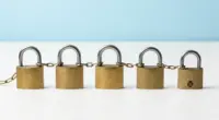 Five brass padlocks in a row on a bright white desk, joined by one continuous chain, the last one open