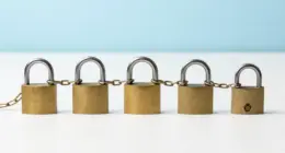Five brass padlocks in a row on a bright white desk, joined by one continuous chain, the last one open