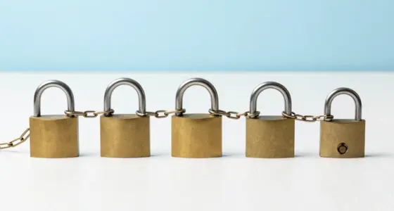 Five brass padlocks in a row on a bright white desk, joined by one continuous chain, the last one open
