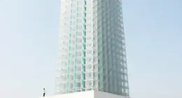 A tall tower of interlocking translucent glass blocks with turquoise light in the joints rises into a pale blue sky, a small person standing at its base