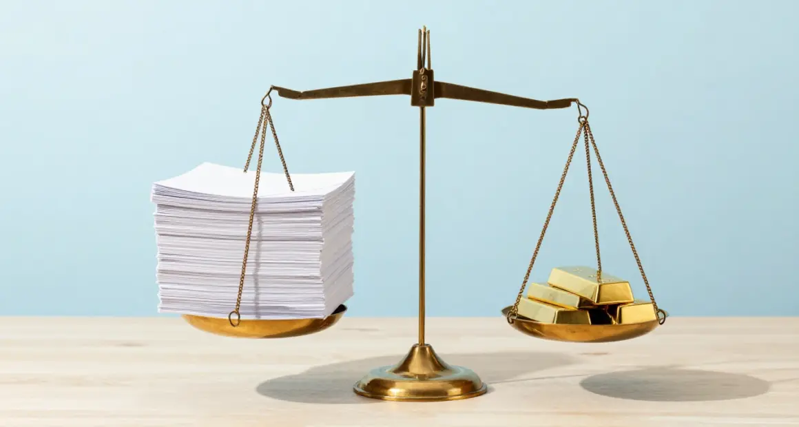 A brass balance scale on a light wooden desk, a tall stack of white documents on one side outweighing a few gold bars on the other