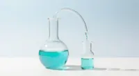 A glass distillation apparatus on a white desk, turquoise liquid dripping from a large flask into a small one