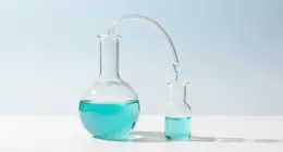 A glass distillation apparatus on a white desk, turquoise liquid dripping from a large flask into a small one