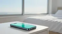 A smartphone glowing softly in pale turquoise on a white bedside table, an empty made bed and a bright window with a pale blue morning sky behind it