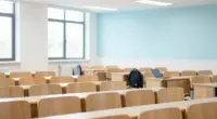 A bright university lecture hall with rows of mostly empty wooden seats, a laptop and a backpack left on one desk
