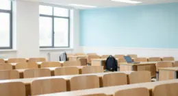 A bright university lecture hall with rows of mostly empty wooden seats, a laptop and a backpack left on one desk