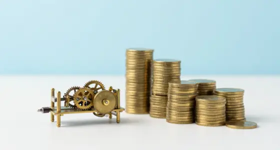 A small brass clockwork engine with gears on a white desk beside tall stacks of plain gold coins
