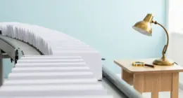 A long line of white paper folders on a conveyor belt leading to a small wooden desk with a brass lamp and a magnifying glass