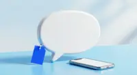 A glossy white speech bubble sculpture on a pale blue desk with a small blank cobalt blue price tag hanging from it, a smartphone lying beside it