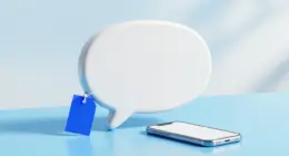 A glossy white speech bubble sculpture on a pale blue desk with a small blank cobalt blue price tag hanging from it, a smartphone lying beside it