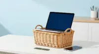 A wicker basket on a bright white table holding an open laptop, with a smartphone and a green memory module lying beside it