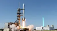 A stainless steel rocket lifting off from a launch tower, with a white data center campus and cooling towers in the background and a turquoise beam of light rising from it