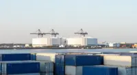 Stacks of blue and white shipping containers in a harbor in the foreground, large white industrial halls with port cranes behind them across calm water