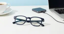 A pair of dark glasses on a bright white desk, a small dark puck-shaped device with a cable, a laptop and a coffee cup behind them